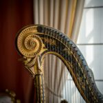 Part of a curved, ornate, gold harp in front of a cream colored curtain and dark red wall