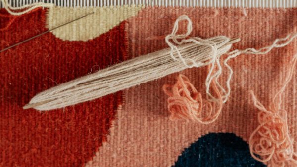 A colorful woven tapestry on a loom with yarn shuttle draped on top