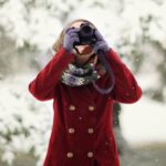 A woman in a red coat aims her camera while snow falls on the trees behind her