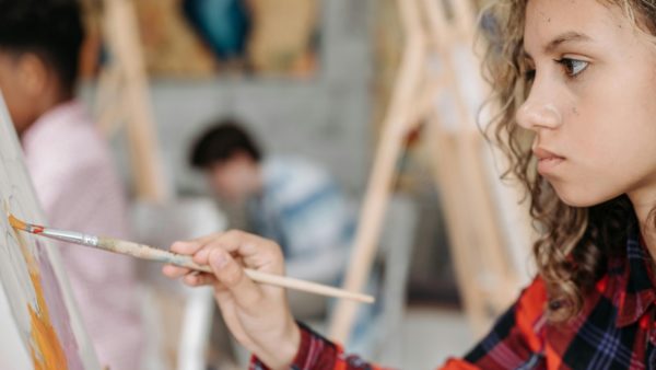 Teenage student painting on a canvas with a long paint brush. They have a serious expression as they concentrate on their work.